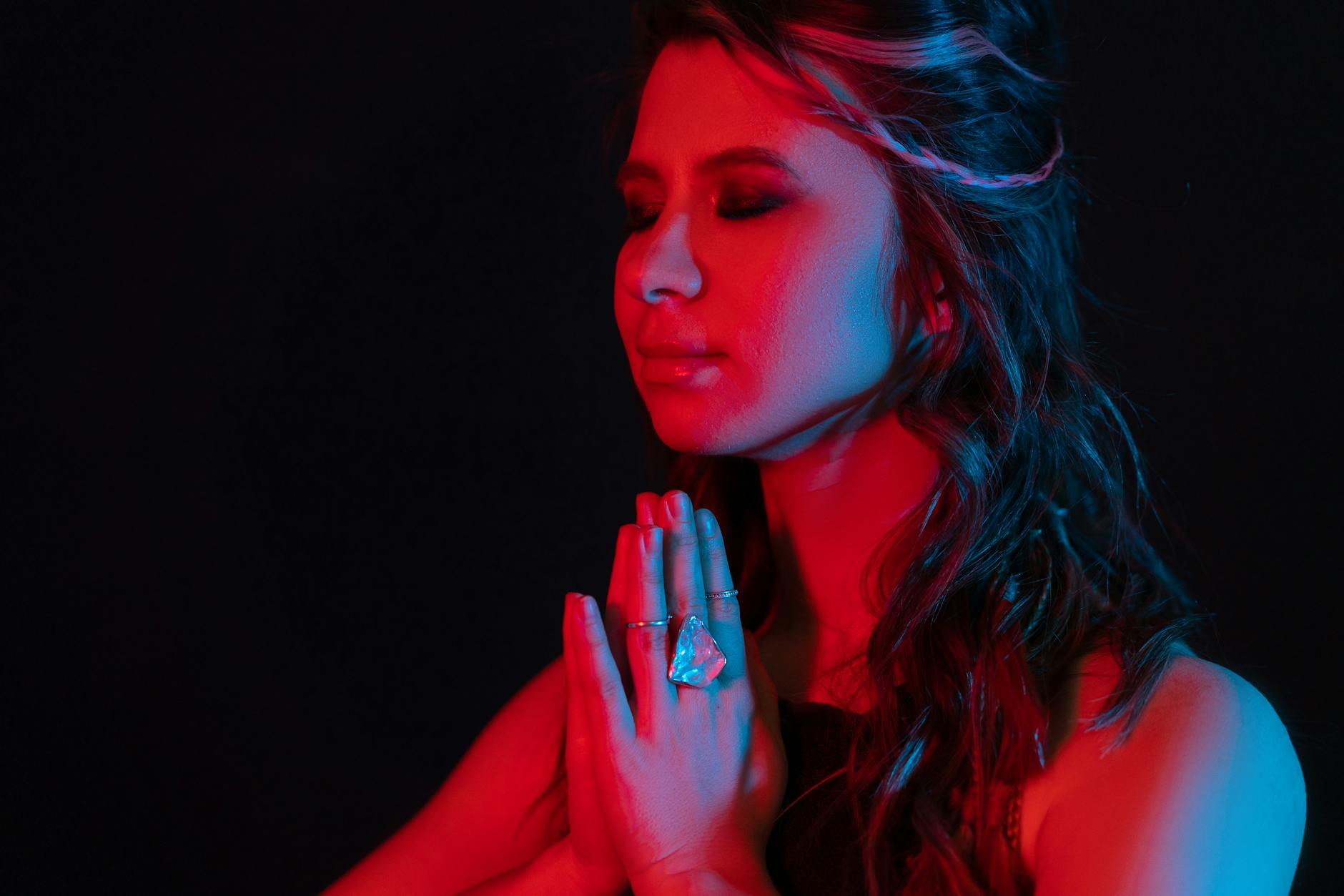 A woman with long hair, dressed in dark clothing, is shown in a contemplative pose with her hands clasped together in front of her, against a dark background illuminated with red and blue lighting.