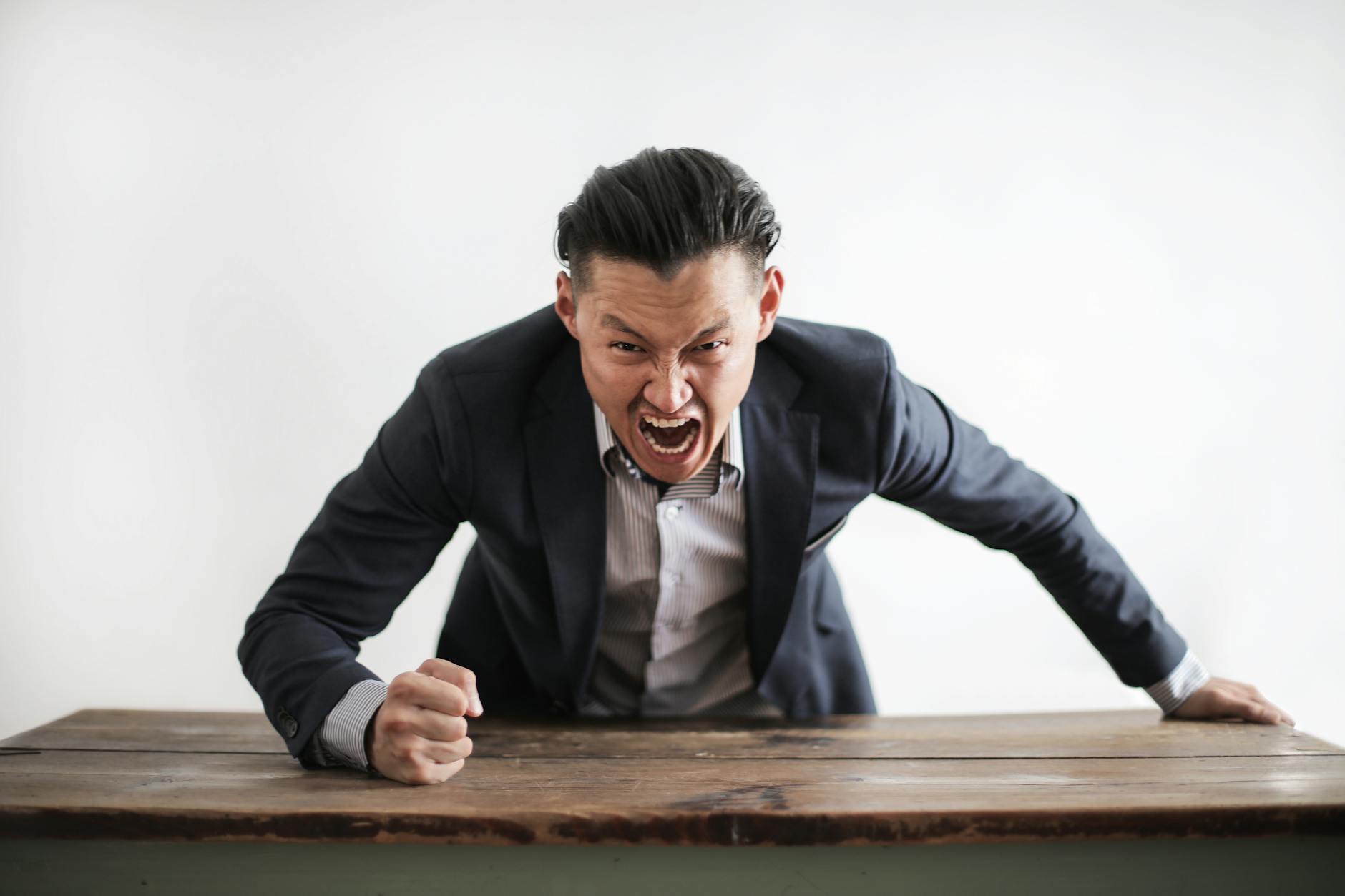 A man in a suit appears to be angrily shouting or expressing frustration while leaning over a wooden table.