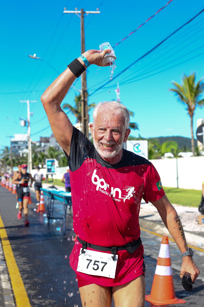 An older man with a full head of gray hair is pouring water from a cup over his head while running in a sunny outdoor setting. He wears a bright red athletic shirt and a race bib number 757. In the background, other runners can be seen along a palm tree-lined street.