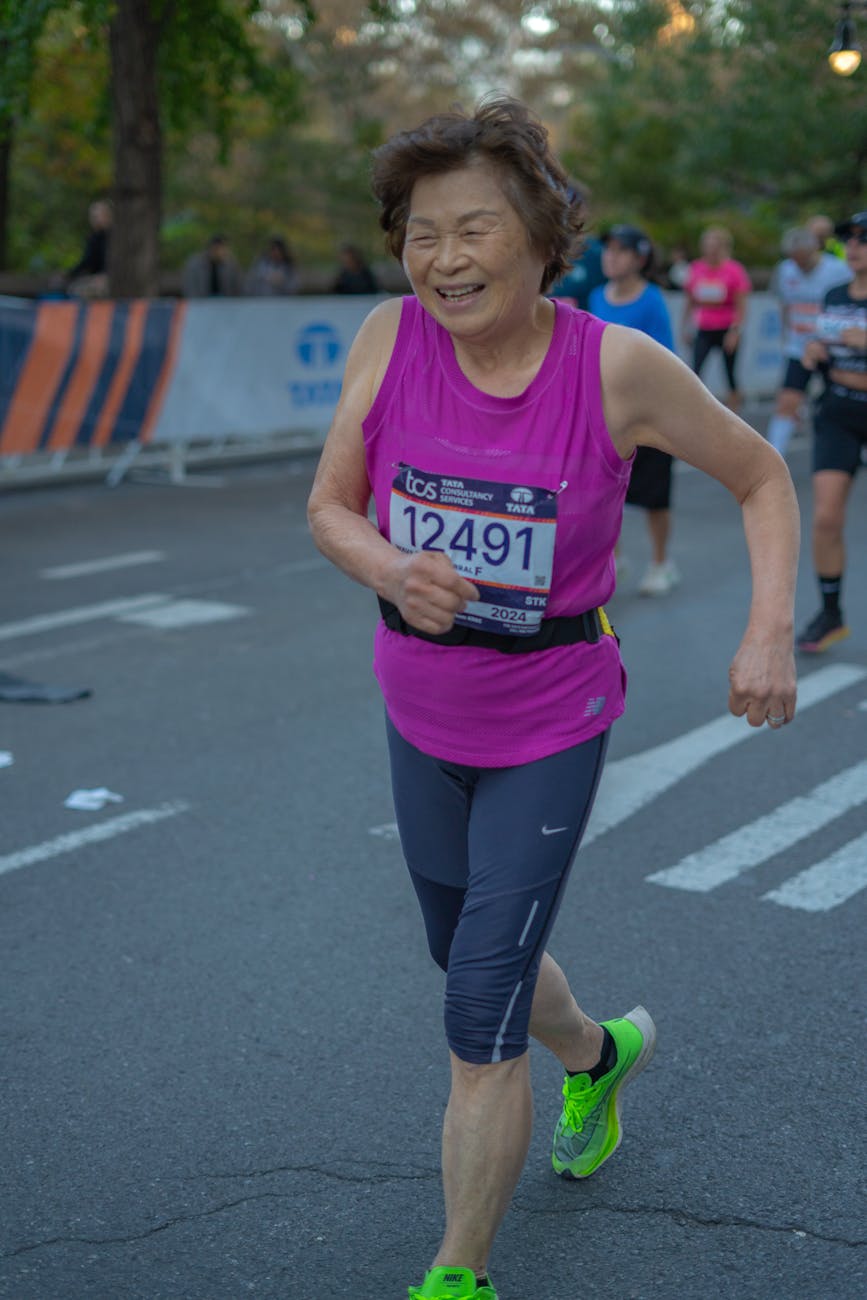 An older female runner smiling while participating in a race, wearing a bright pink tank top and bib number 12491, with greenery and other runners in the background.