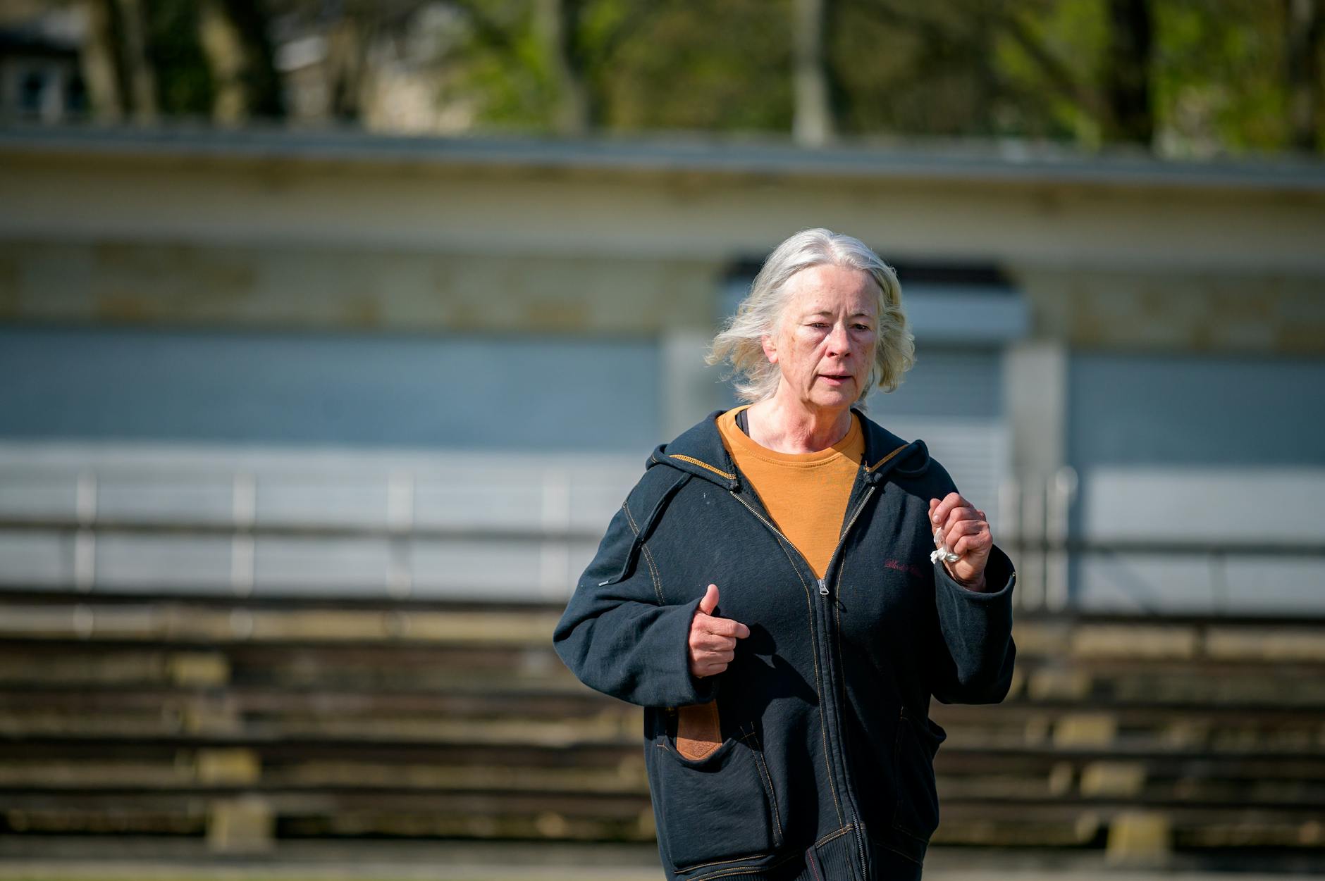 An older woman running outdoors in a black hoodie and orange shirt, focused on her exercise, with a blurred background of benches and greenery.