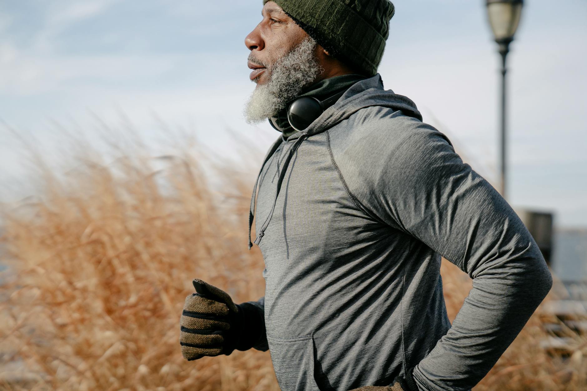 Older man jogging outdoors in a gray hoodie and gloves, surrounded by tall grass with a calm sky in the background.
