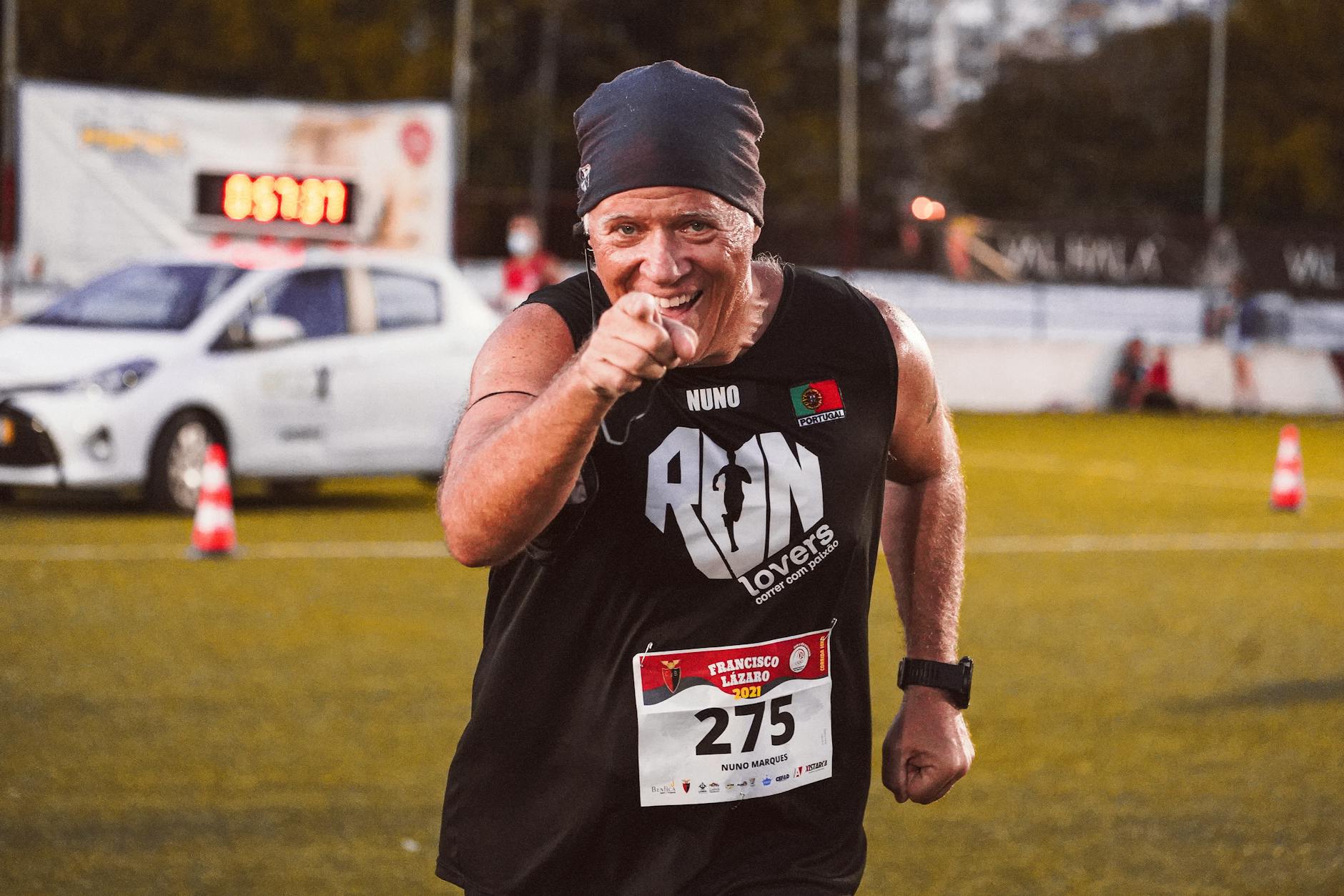 A runner with a confident expression points at the camera while sporting a black sleeveless shirt with 'RUN lovers' printed on it, a race bib, and a headband, during an event in a sporting field.