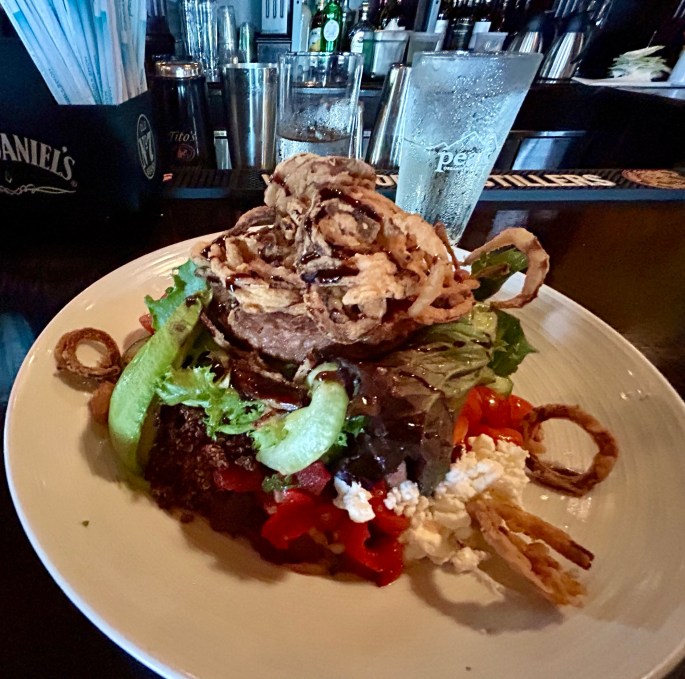 A colorful salad topped with a fried onion ring stack, featuring greens, tomatoes, and cheese, served at a bar setting.