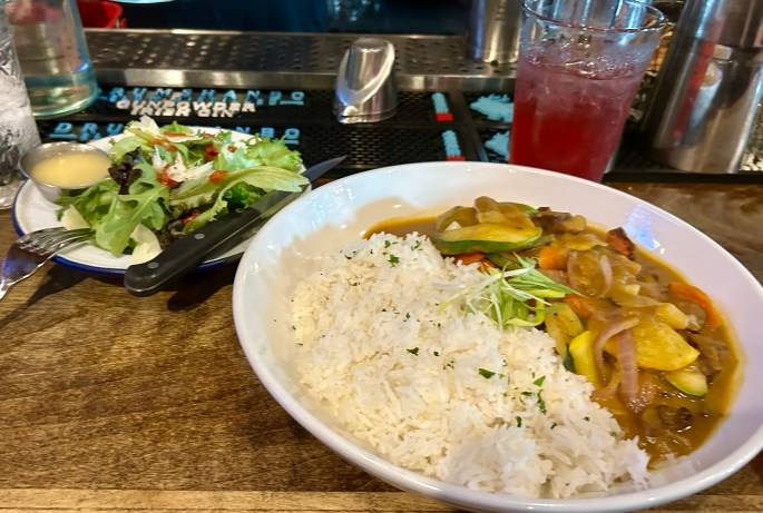 A plate of rice accompanied by a colorful vegetable curry, served with a side salad and a drink in the background.