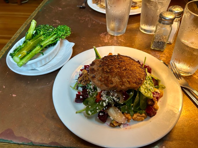 A plate of salad with mixed greens, nuts, and beets topped with a vegetable patty, accompanied by a side of steamed broccoli.