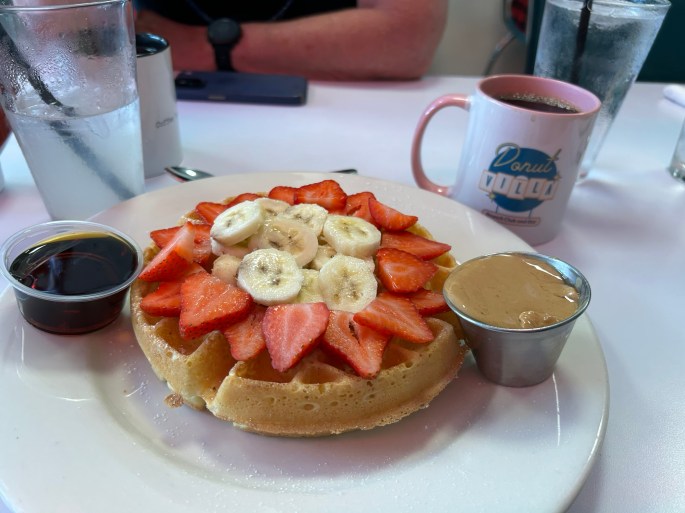 A plate with a waffle topped with sliced strawberries and bananas, accompanied by a small cup of peanut butter and a small container of syrup, set on a table with beverages in the background.
