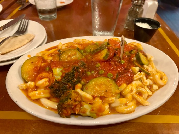 A plate of pasta topped with marinara sauce and mixed vegetables, including zucchini, broccoli, and peas, served in a restaurant setting.