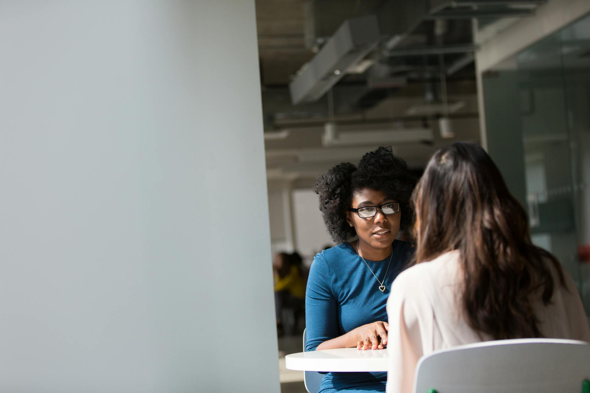 Two women engaged in a conversation at a table in a modern office setting, with one woman wearing glasses and a blue dress, and the other facing away.