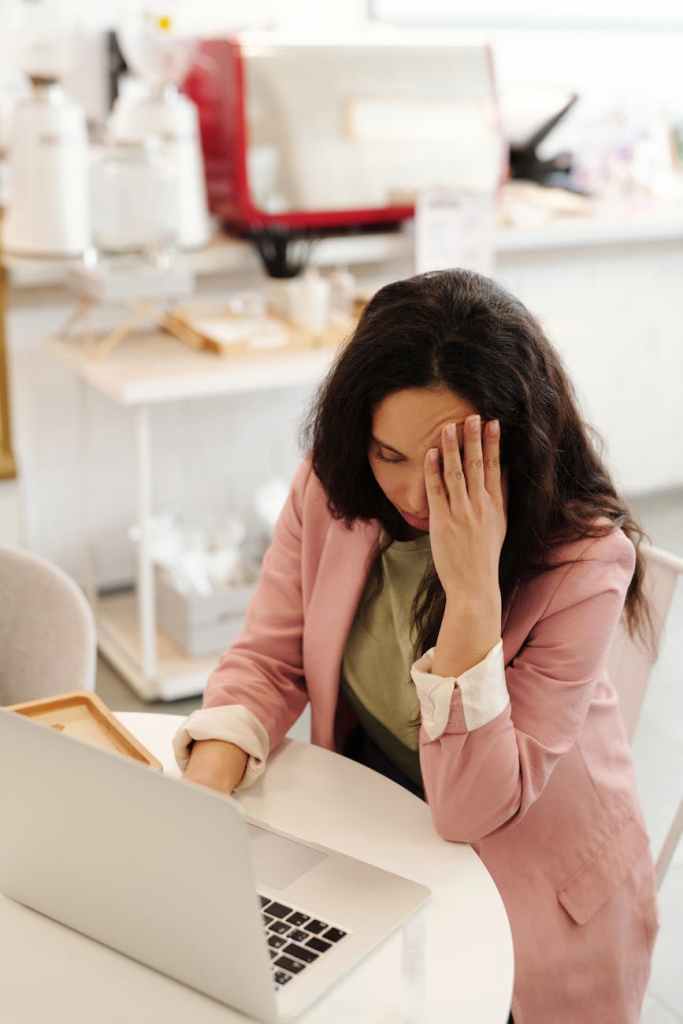 A woman in a pink blazer sitting at a table, looking stressed or overwhelmed while using a laptop.