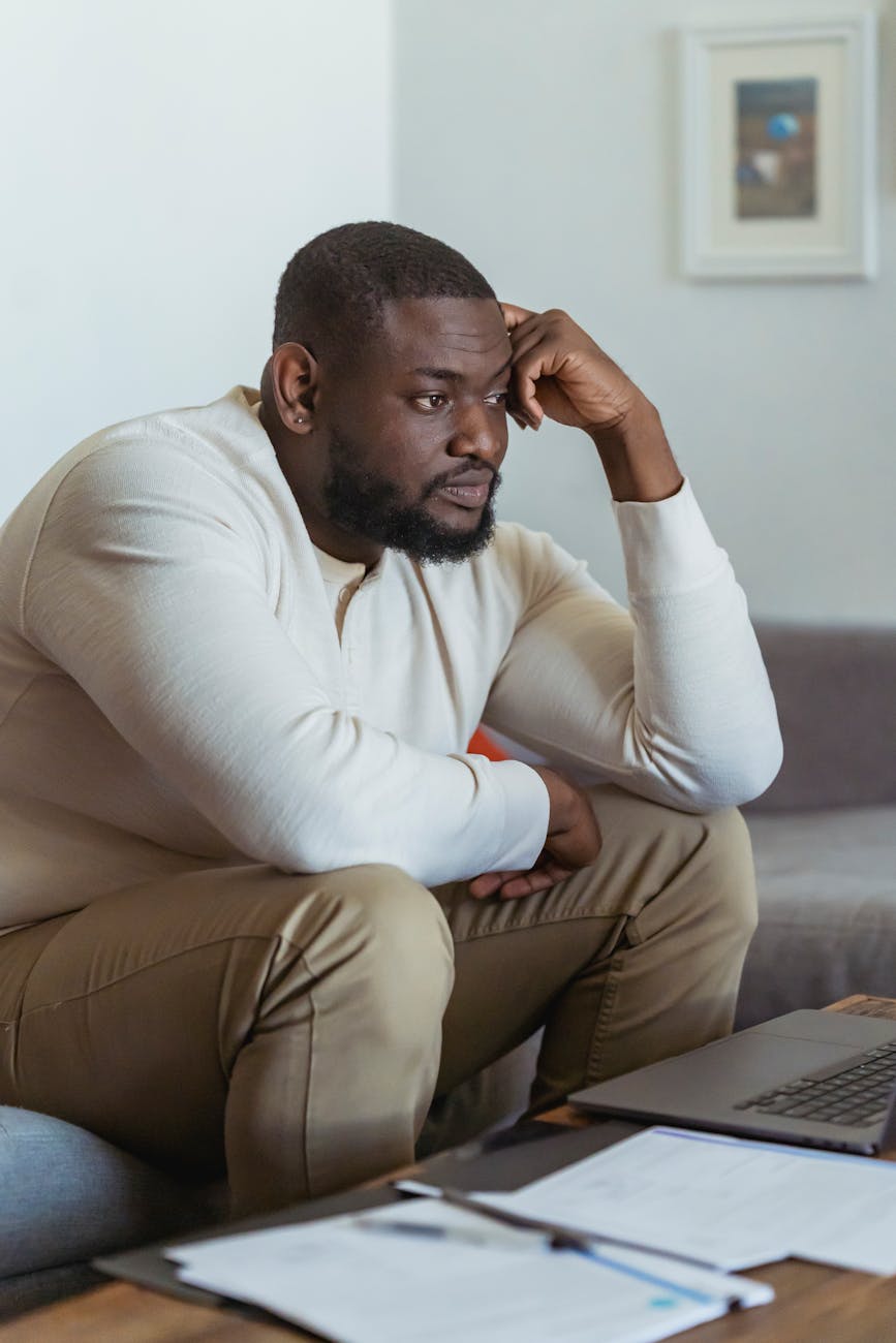 A person sitting on a couch, resting their chin on their hand, looking thoughtfully at a laptop with papers and a pen on the table.