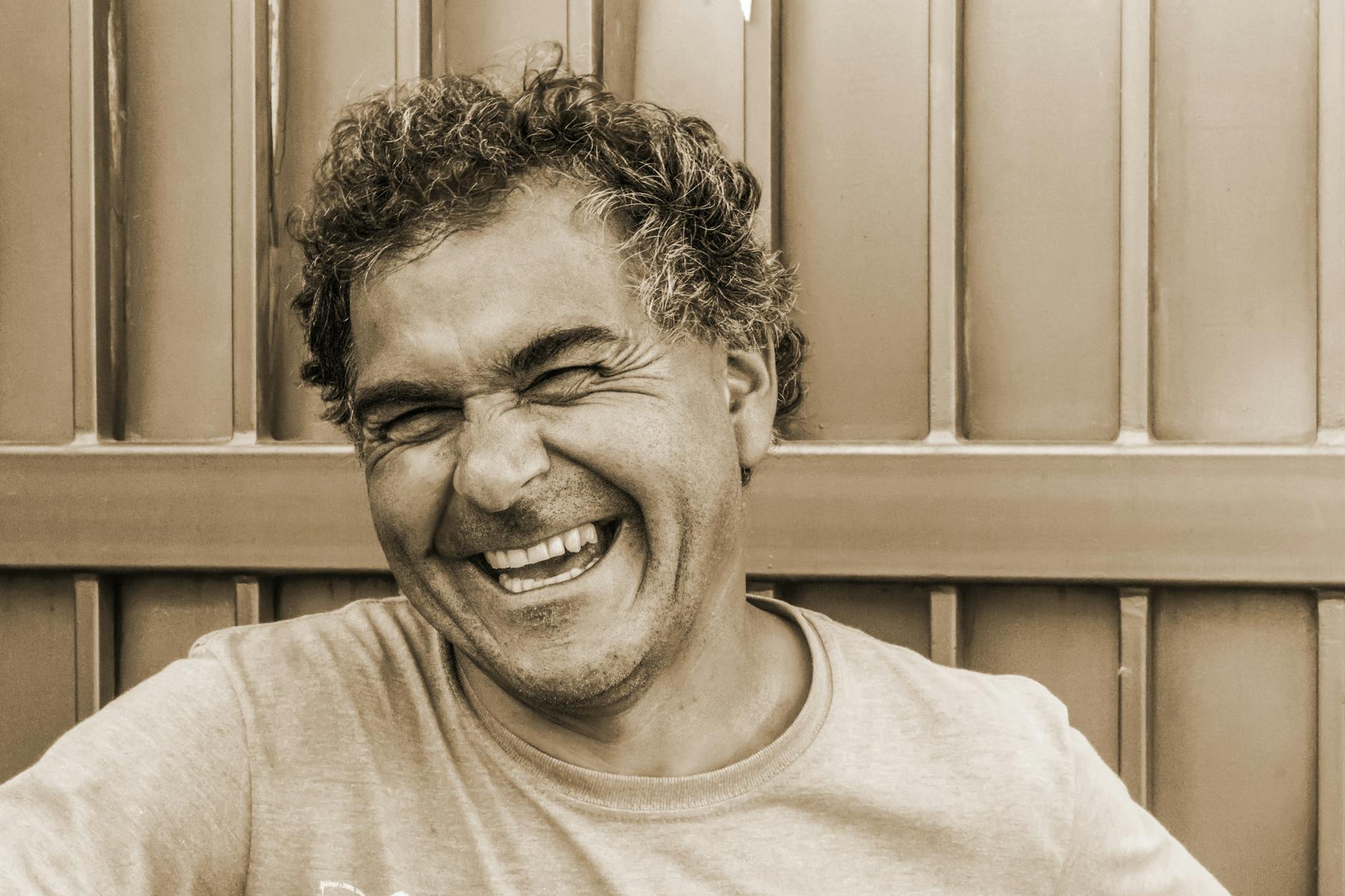 A smiling man with curly hair, laughing joyfully against a wooden background.