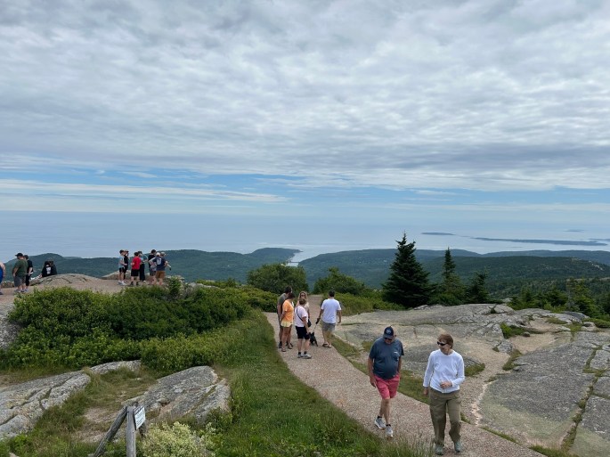 A group of hikers enjoying the scenic views from the summit of Cadillac Mountain in Acadia National Park, with lush greenery and ocean in the background under a cloudy sky.