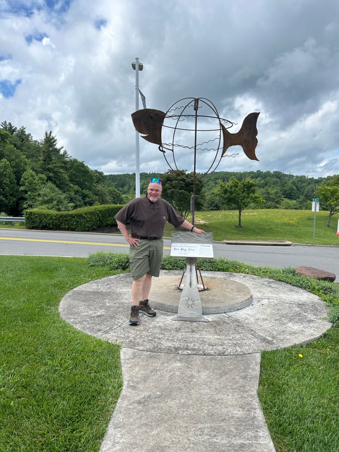 A man stands next to a large fish sculpture on a circular concrete platform, surrounded by grass and trees, under a cloudy sky.