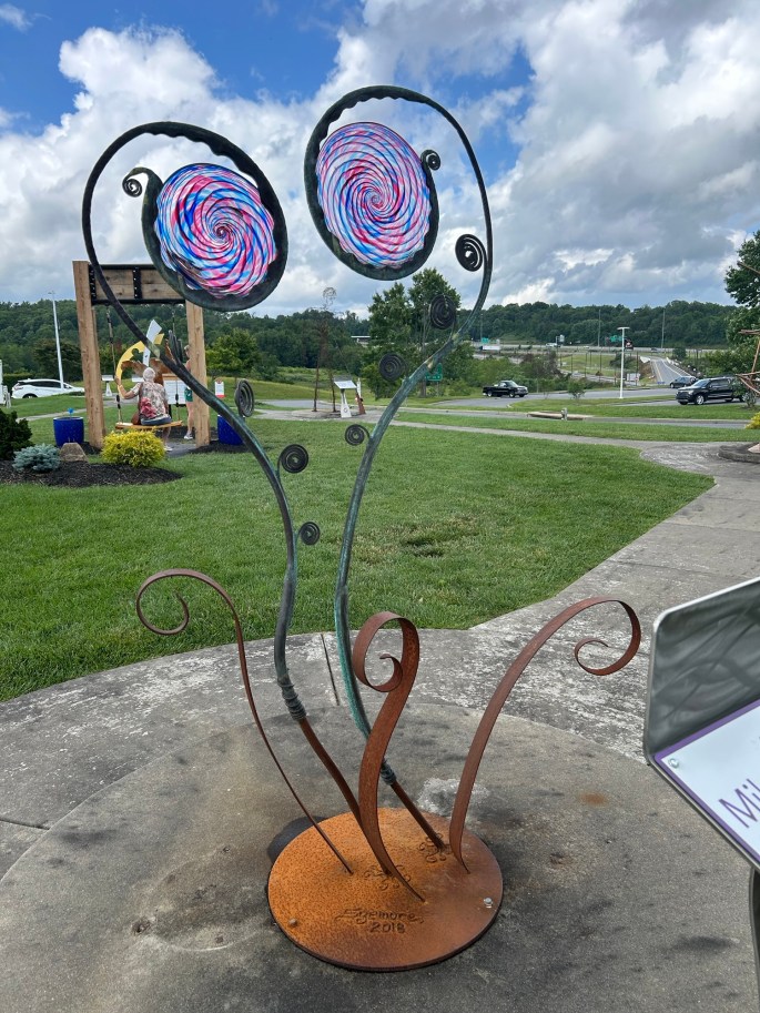 A colorful outdoor metal sculpture featuring two spiral-shaped elements atop slender, twisting stems, set against a grassy landscape and cloudy sky.