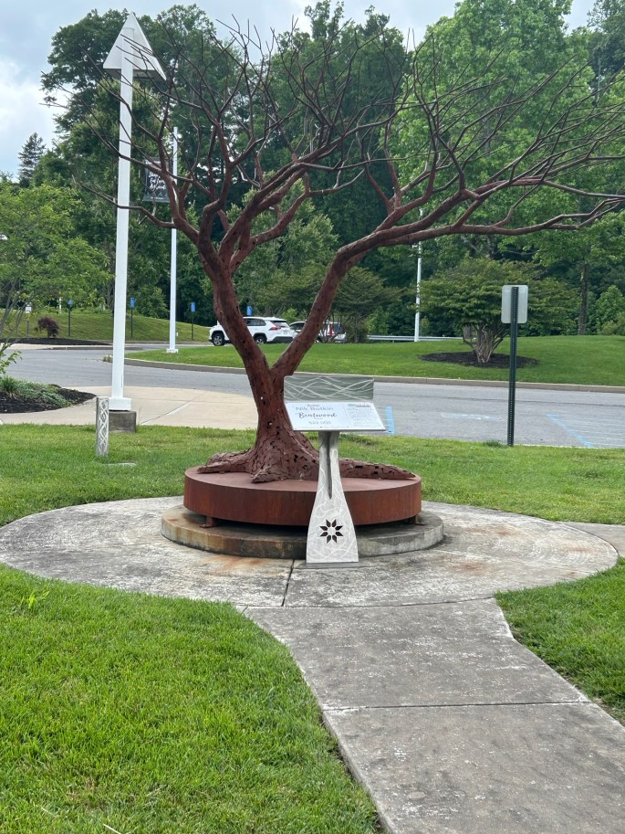 A sculptural tree installation with bare branches, situated on a circular stone base, with a descriptive plaque nearby.