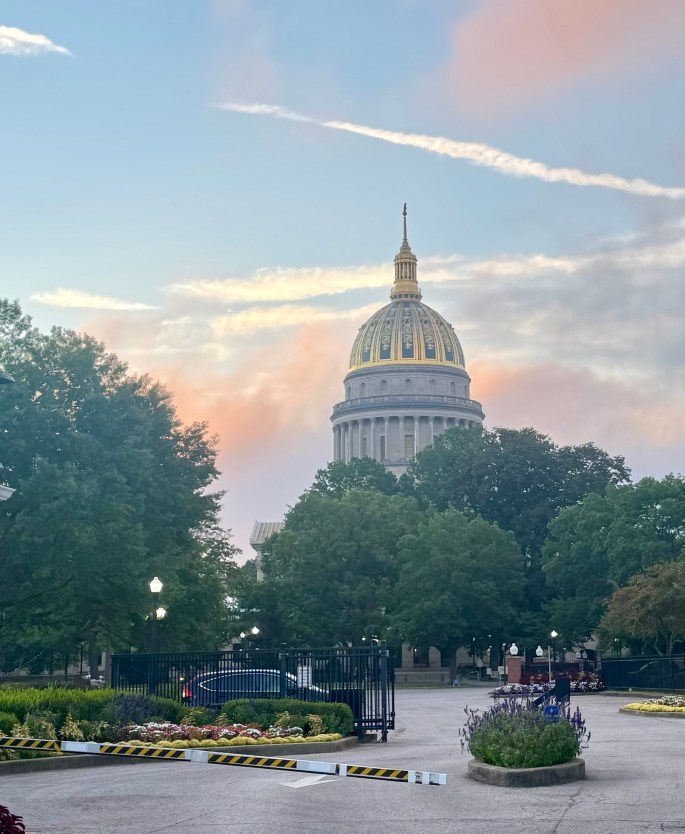 The West Virginia State Capitol building with a golden dome, surrounded by trees and a pastel sky at dawn.