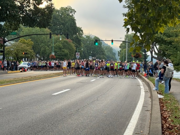 Runners lined up at the starting line of a race on a road, with traffic lights and spectators in the background.