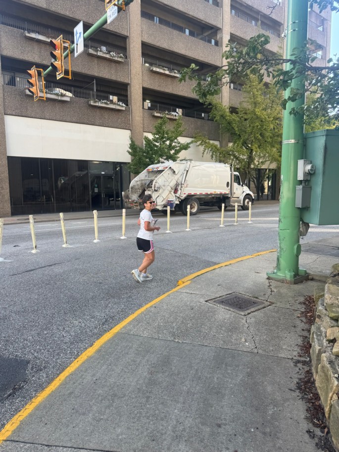 A person jogging on a city street with a garbage truck parked nearby and a multi-story building in the background.