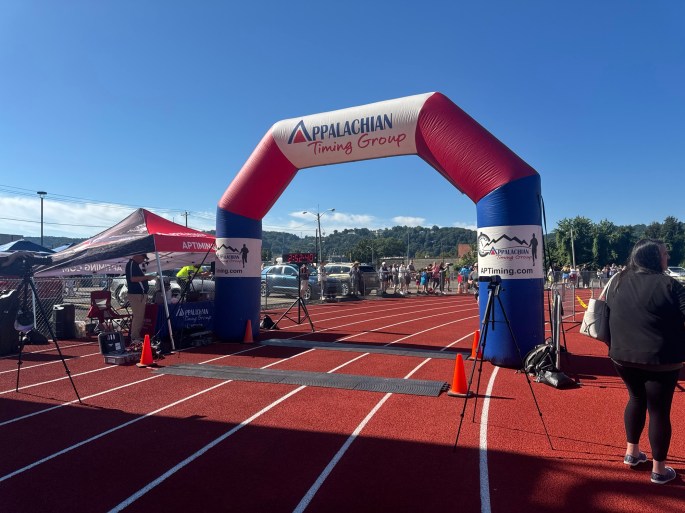 A running event starting line with an inflatable arch labeled 'Appalachian Timing Group' on a red track, set against a clear blue sky and distant hills.