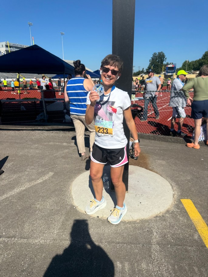 A smiling woman holding a medal, wearing athletic clothing, stands next to a post in a race finish area, with a crowd and tents in the background.