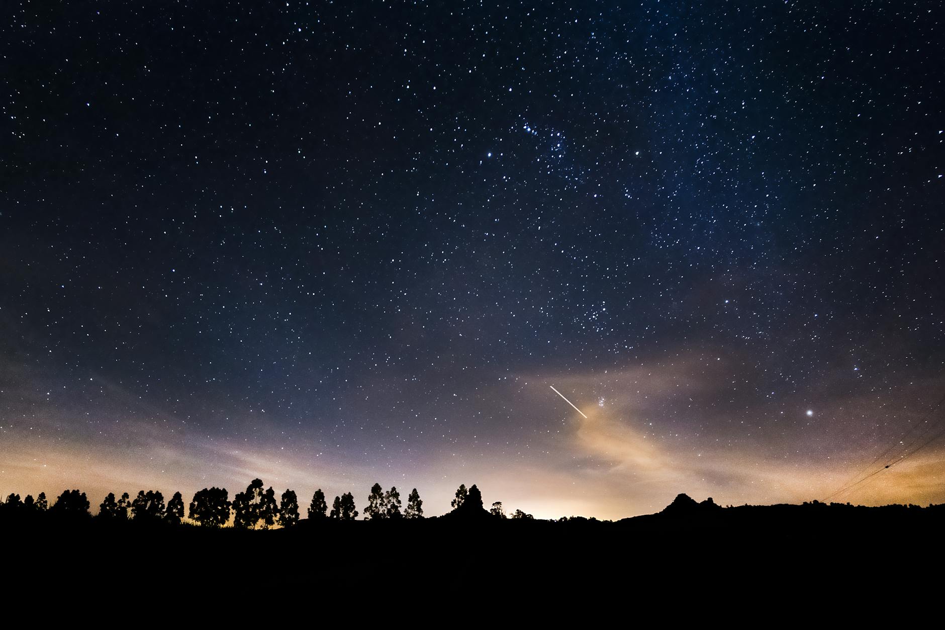 A night sky filled with stars, with a shooting star streaking across the horizon, silhouetted by trees and distant hills.