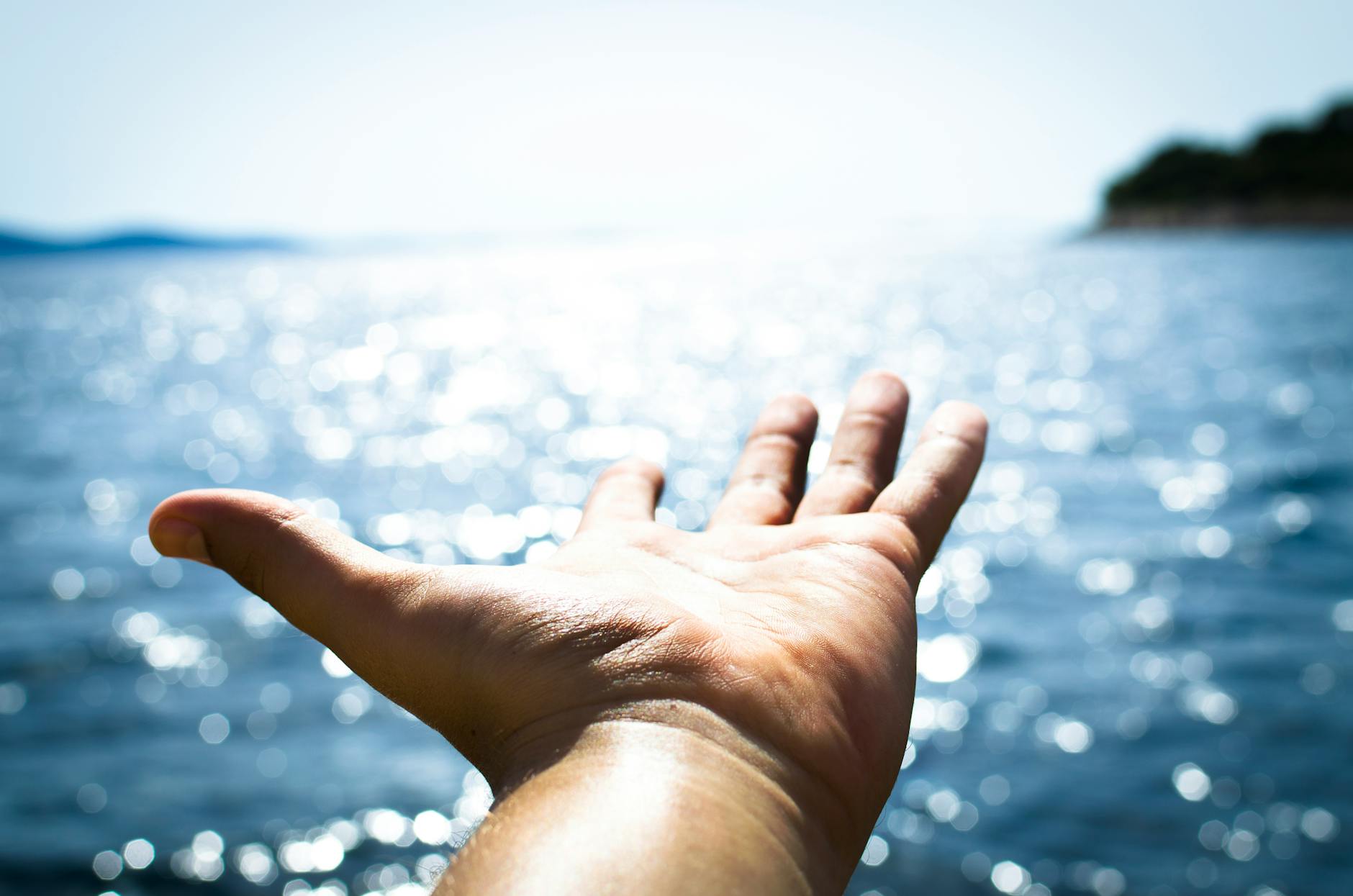 A hand reaching out towards a shimmering body of water under a bright sky.