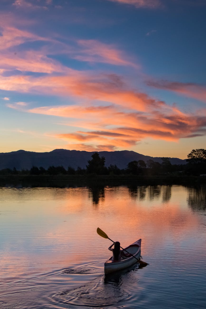 A person kayaking on a calm body of water during sunset, with vibrant pink and orange clouds reflected in the water.