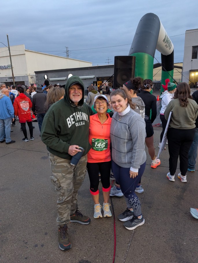 A group of three people, including a man in a green hoodie, a woman in a bright orange top with a running bib, and a woman in a gray sweater, gather smiling at a running event in an outdoor setting with other runners in the background.
