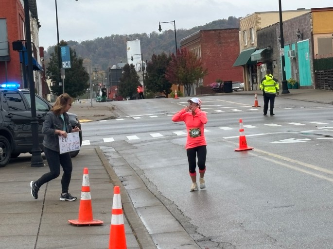 A female runner in a bright pink shirt crosses a street marked by traffic cones, while a supporter holds a sign nearby in a lively downtown setting.
