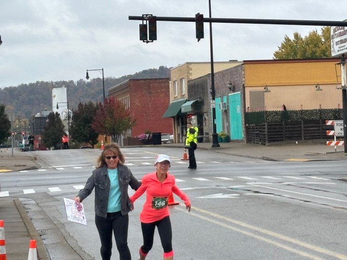 Two women running together on a city street during a marathon, with one holding a sign. They are surrounded by a lively atmosphere and shops in the background.