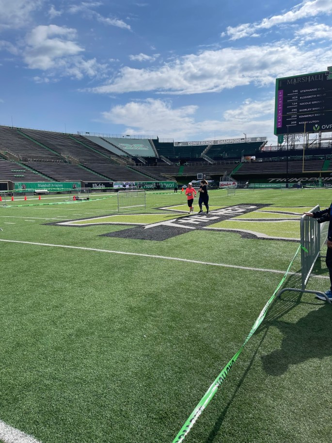 A runner crosses the finish line at a sports stadium, with spectators in the stands and a clear blue sky overhead.