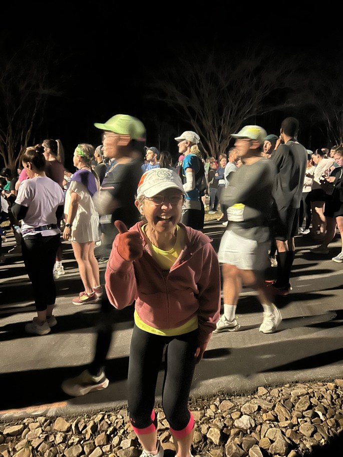 A smiling woman with glasses and a cap gives a thumbs-up gesture in a crowd of runners at night, with other participants blurred in motion around her.
