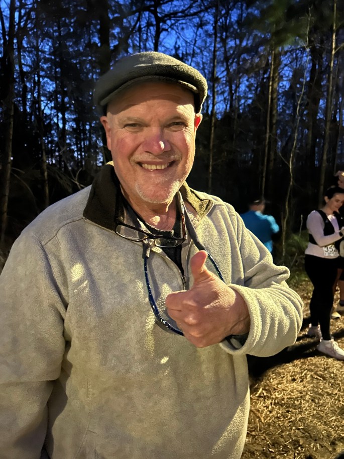 A smiling older man wearing a cap and light-colored jacket gives a thumbs-up gesture outdoors, surrounded by trees during dusk.
