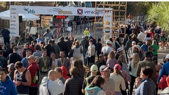 A large crowd of participants and spectators gathered at an outdoor event, with banners and a timer displaying '2:26:06' in the background, near a finish line.
