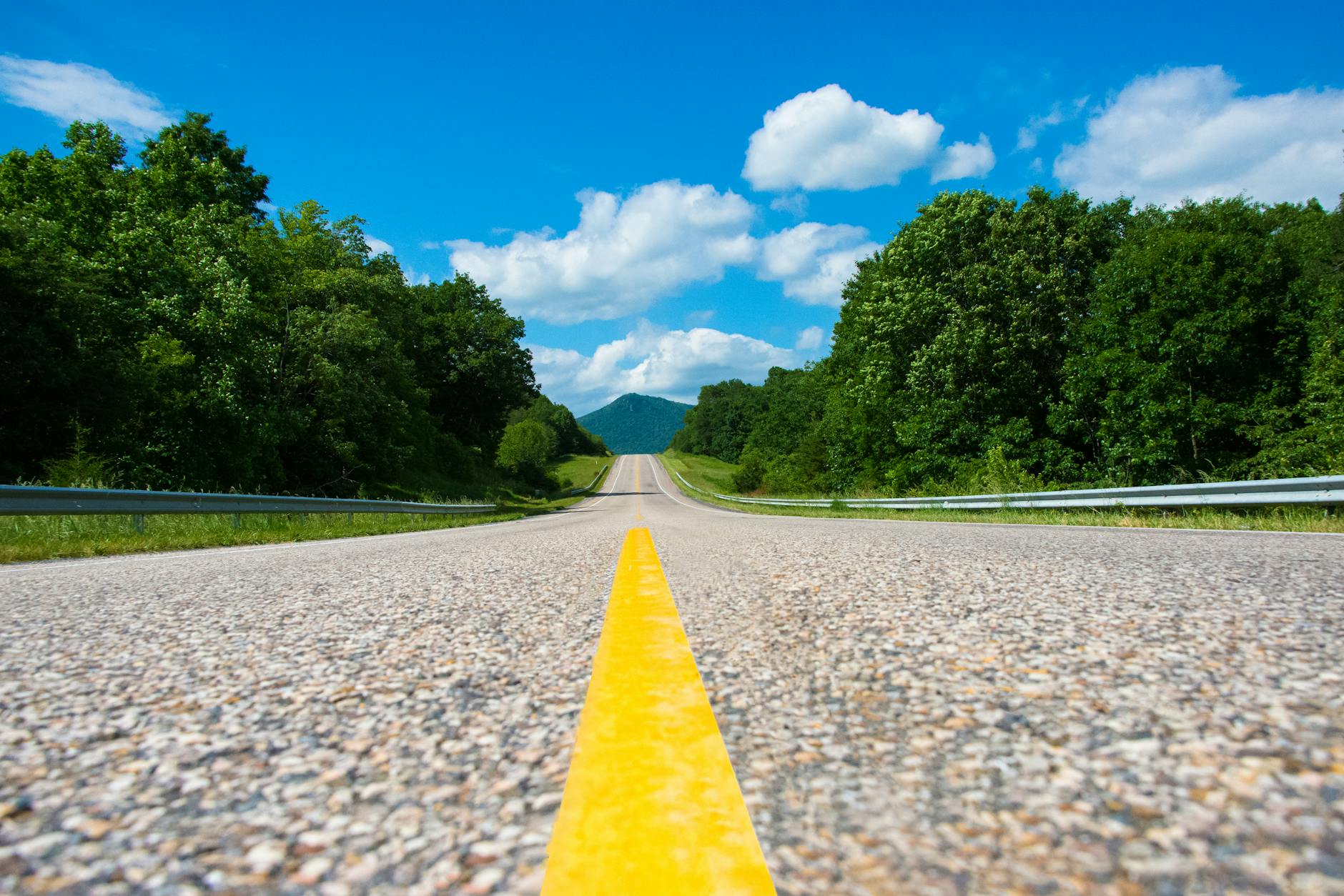 A winding asphalt road stretches into the distance, flanked by lush green trees under a bright blue sky with fluffy white clouds.