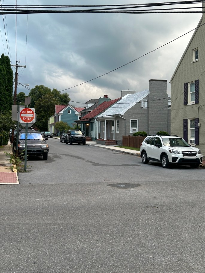 A quiet street scene with several houses on either side, featuring a mix of architectural styles. Parked cars line the road, and a 'Do Not Enter' sign is visible in the foreground. The sky is cloudy, suggesting overcast weather.
