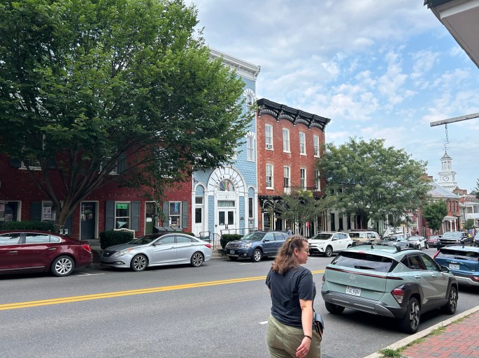 A street scene featuring a woman walking alongside parked cars and historic buildings with varying architectural styles under a partly cloudy sky.