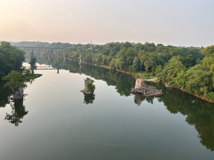 A calm river reflecting the surrounding trees and an old railway bridge in the background, with small rocky outcrops emerging from the water.