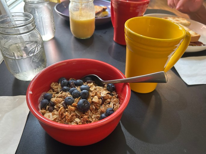 A red bowl filled with granola and fresh blueberries, accompanied by a yellow mug, glass of water, and other breakfast items in the background.